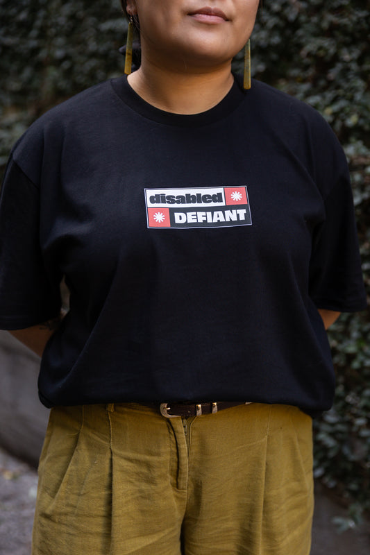 Eda wearing a black T-shirt with "disabled, DEFIANT" in bold text stands outdoors. The design is black, red and white, and features two tohuwhetū.