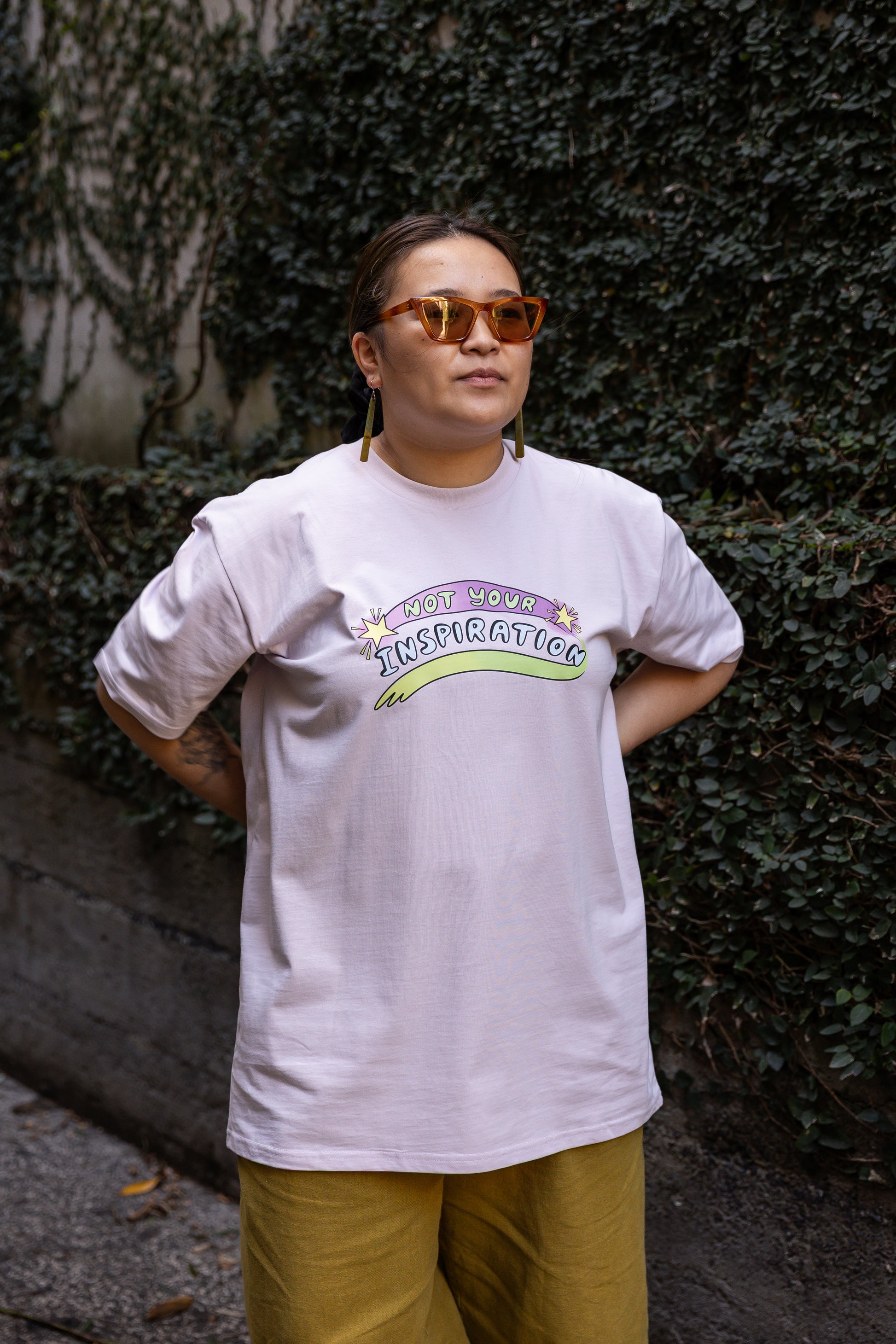 Eda wearing a light pink T-shirt with the phrase "Not Your Inspiration" in colourful letters stands against a leafy background. The words 'Not your' appear in lime green on a purple swoosh, positioned between two yellow stars. Beneath these words, 'inspiration' appears in all caps, while a second stroke of colour, which appears like a green-yellow paint stroke, provides a boundary of support at the base of the print. 