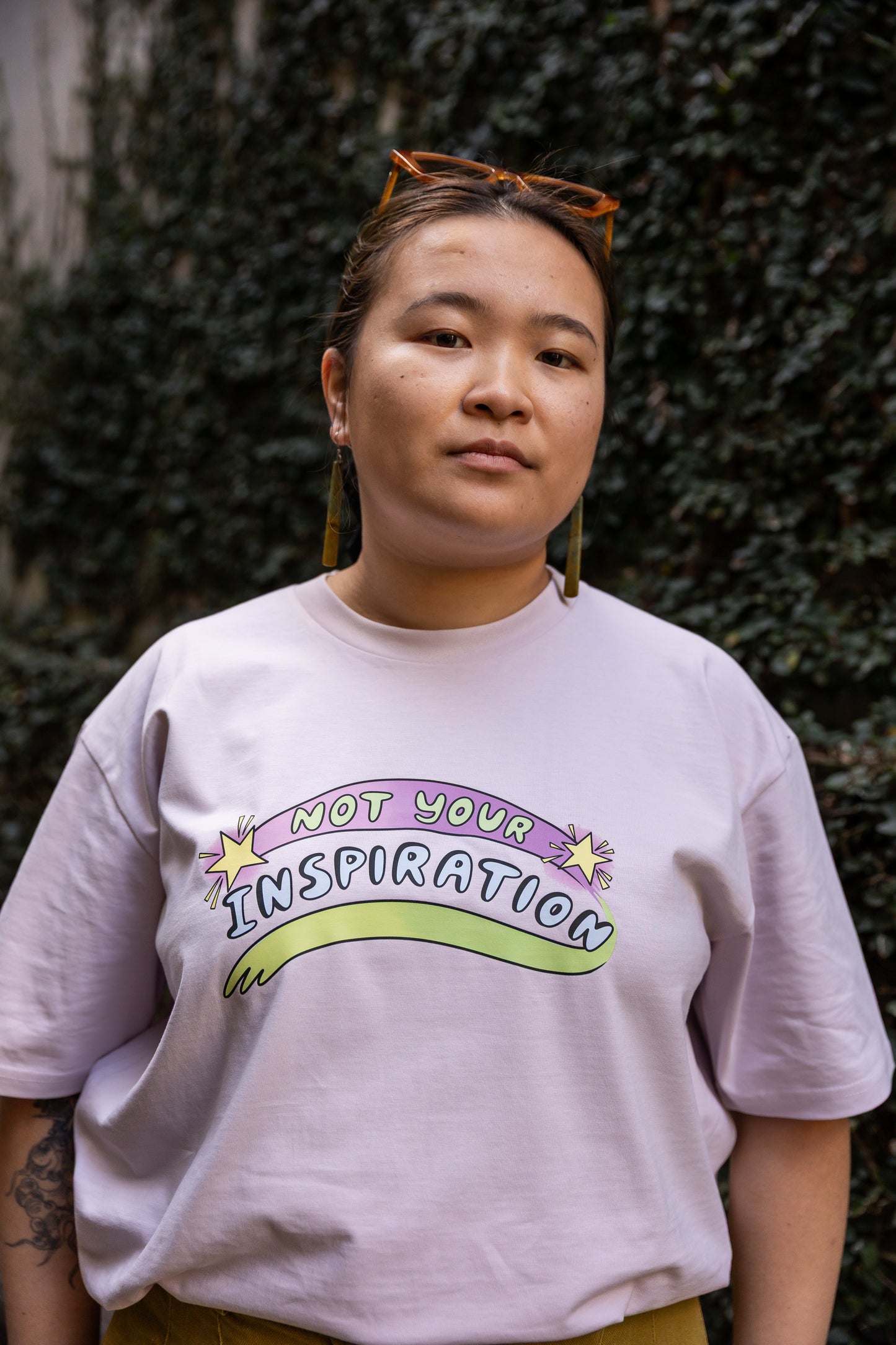 Eda wearing a light pink T-shirt with the phrase "Not Your Inspiration" in colourful letters stands against a leafy background. The words 'Not your' appear in lime green on a purple swoosh, positioned between two yellow stars. Beneath these words, 'inspiration' appears in all caps, while a second stroke of colour, which appears like a green-yellow paint stroke, provides a boundary of support at the base of the print. 