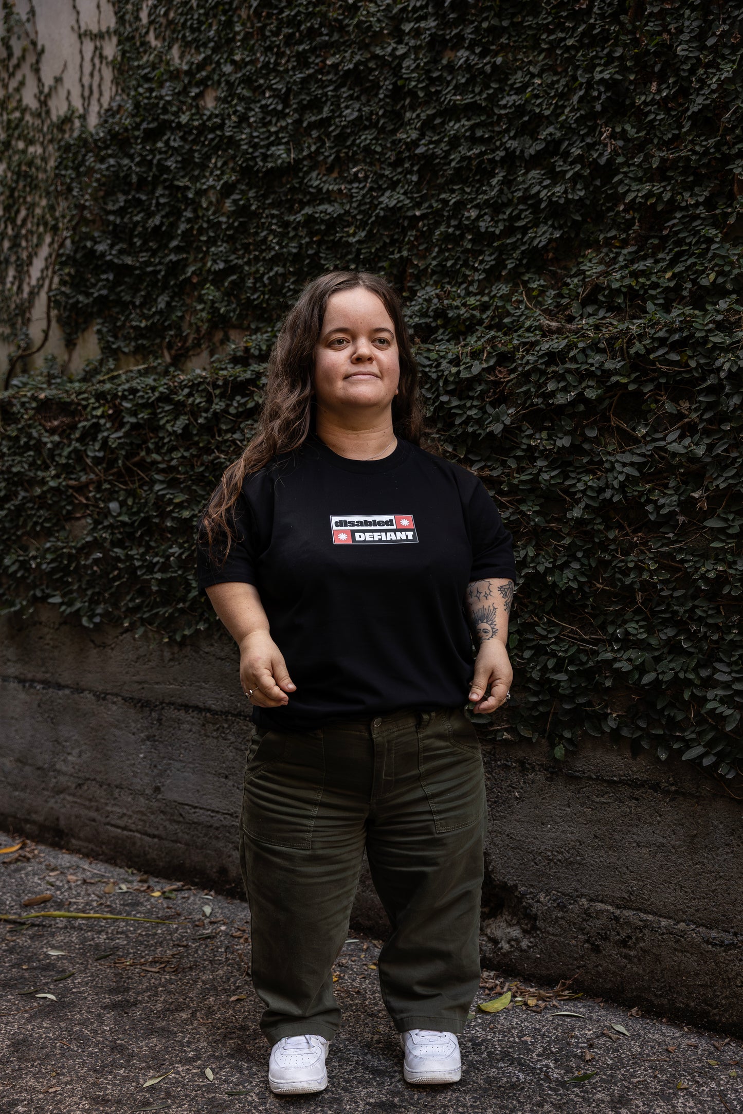 Ella stands in-front of a leafy wall outdoors, wearing a black T-shirt with "disabled, DEFIANT" in bold text stands outdoors. The design is black, red and white, and features two tohuwhetū.