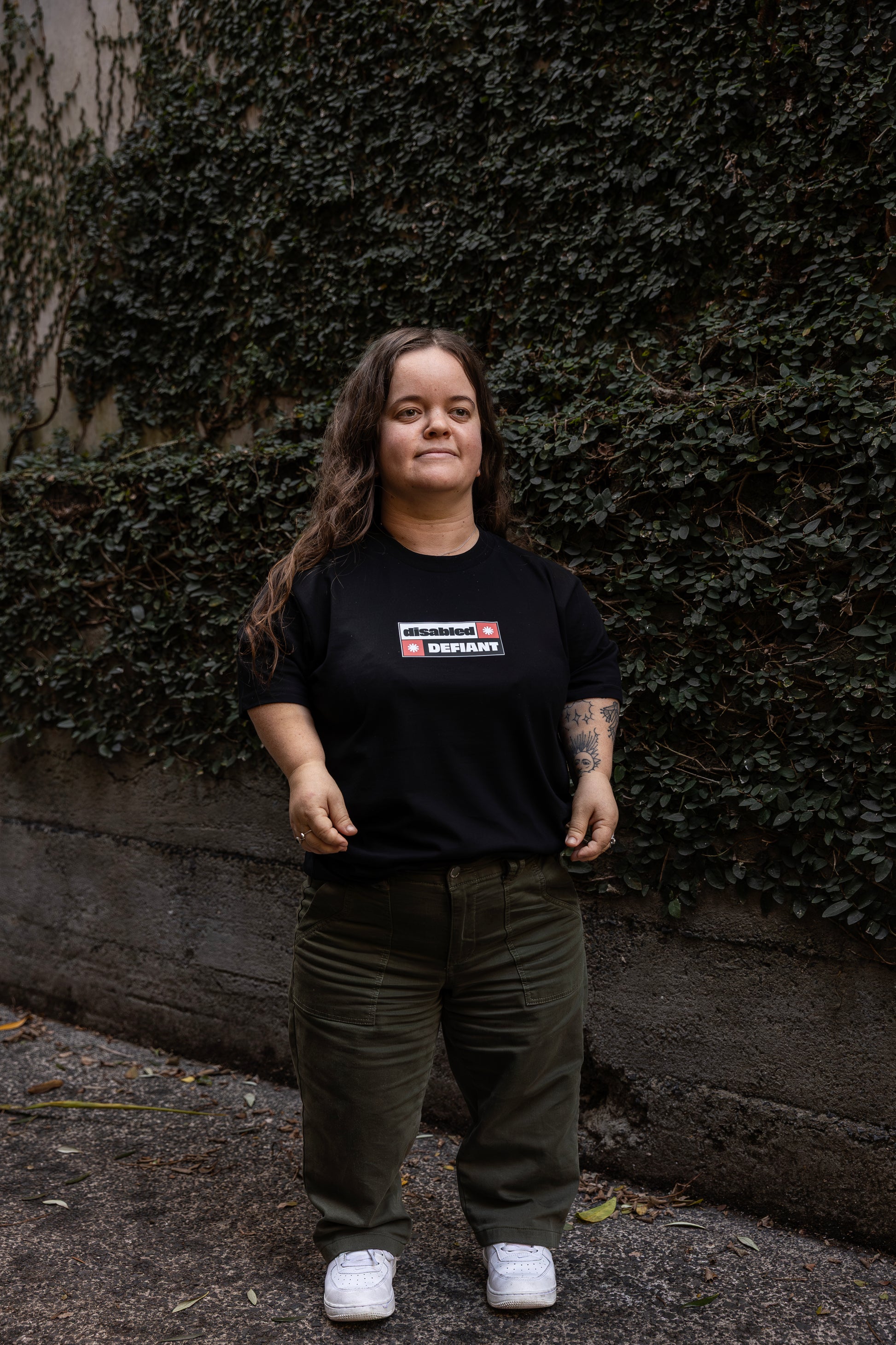 Ella stands in-front of a leafy wall outdoors, wearing a black T-shirt with "disabled, DEFIANT" in bold text stands outdoors. The design is black, red and white, and features two tohuwhetū.