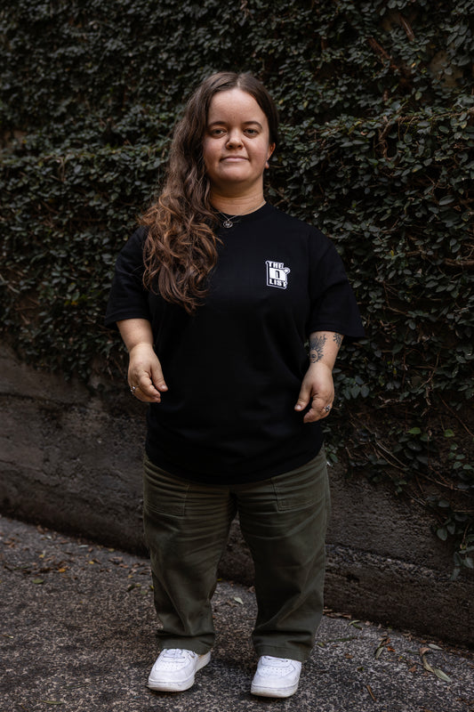 Ella stands outdoors against a wall covered with green ivy. She wears a black T-shirt with a white "The D*List" logo on the left chest, olive pants, and white shoes.