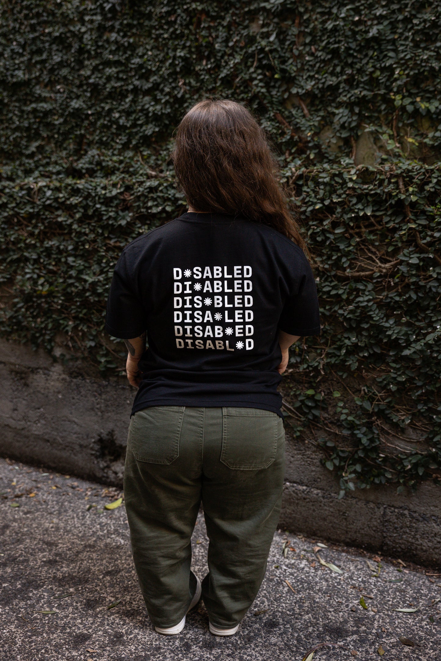 Ella stands facing away from the camera, wearing a black T-shirt with "DISABLED" printed multiple times. The white print on the back reads "DISABLED" repeated six times with a tohuwhetū replacing different letters from left to right on each line. She stands on a leaf-strewn path beside a vine-covered wall.