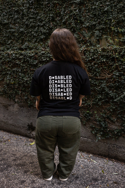 Ella stands facing away from the camera, wearing a black T-shirt with "DISABLED" printed multiple times. The white print on the back reads "DISABLED" repeated six times with a tohuwhetū replacing different letters from left to right on each line. She stands on a leaf-strewn path beside a vine-covered wall.