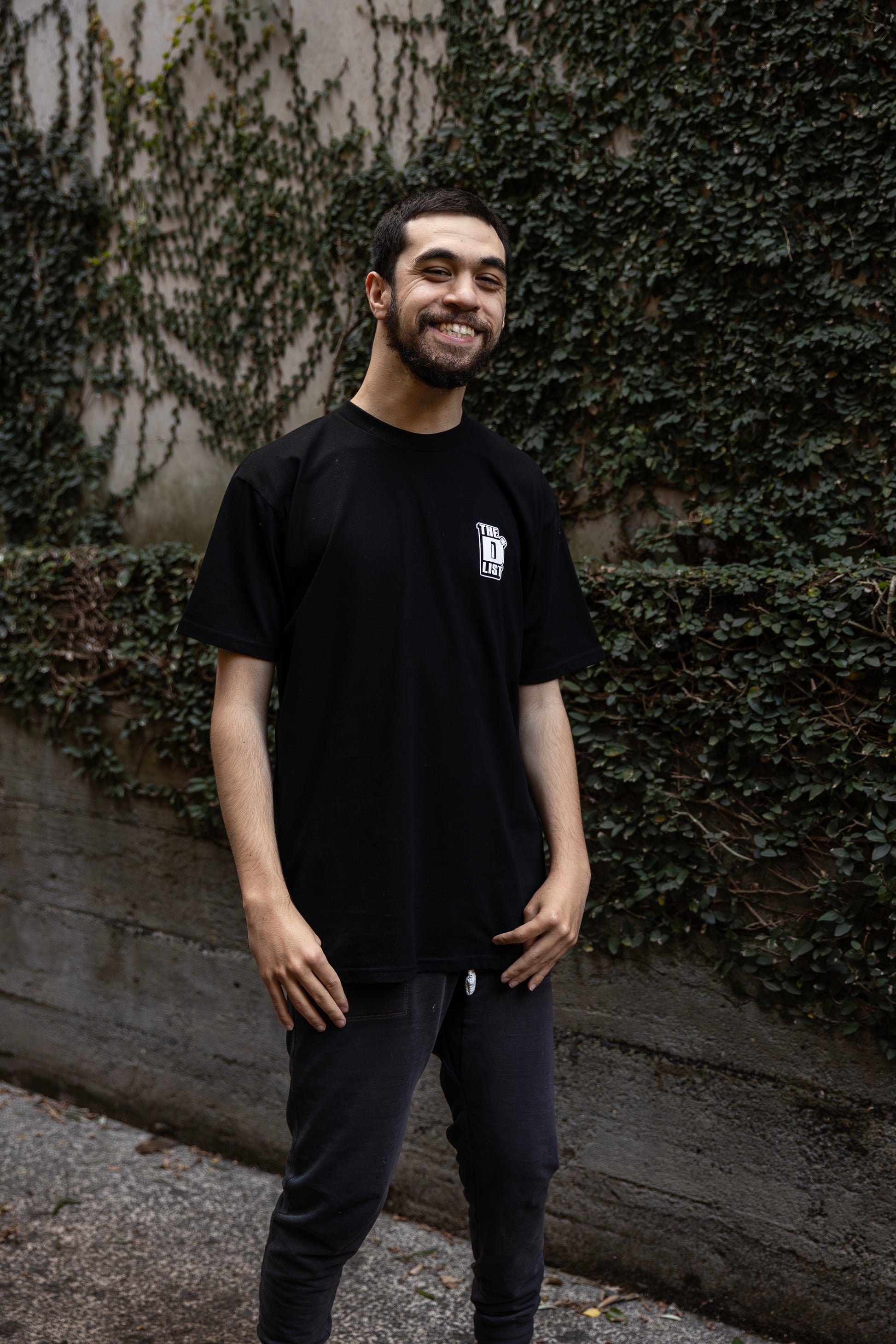 Will wearing a black t-shirt with "The D*List" logo on the left chest stands outdoors, with an ivy-covered wall behind him.