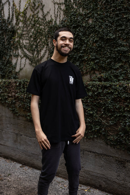 Will wearing a black t-shirt with "The D*List" logo on the left chest stands outdoors, with an ivy-covered wall behind him.