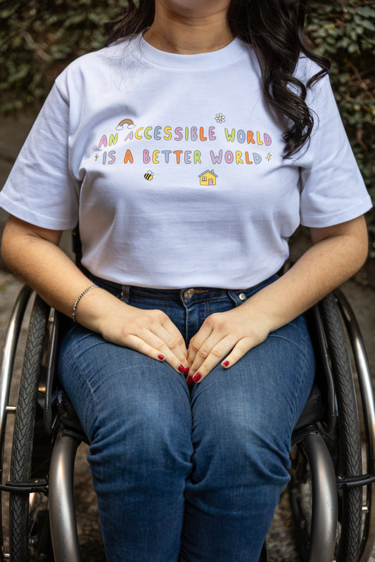 Olivia sits in her wheelchair wearing jeans and a white t-shirt with colourful text which reads "An accessible world is a better world". The design has small designs around it including flowers, a rainbow, a bee, a house and a star.
