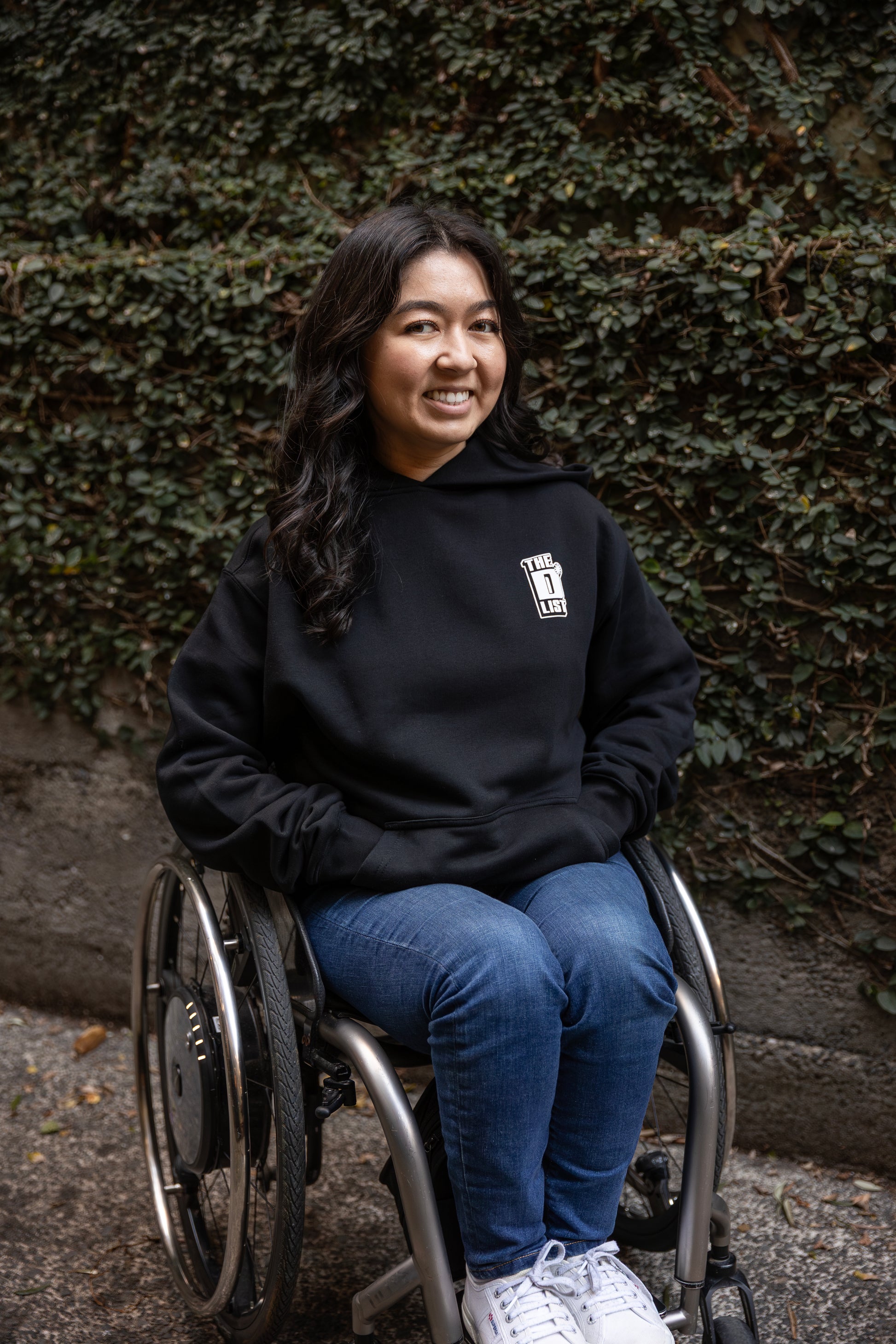 Olivia, who is sitting in a wheelchair, wears a black hoodie with 'The D*List' logo on the left chest, and blue jeans. The background is a wall covered with dense green ivy.