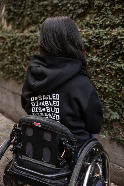 Olivia, sitting in a wheelchair, faces away, wearing a black hoodie with "DISABLED" printed repeatedly in white. Tohuwhetū replace different letters from left to right on each line. Greenery lines the stone path beside them.