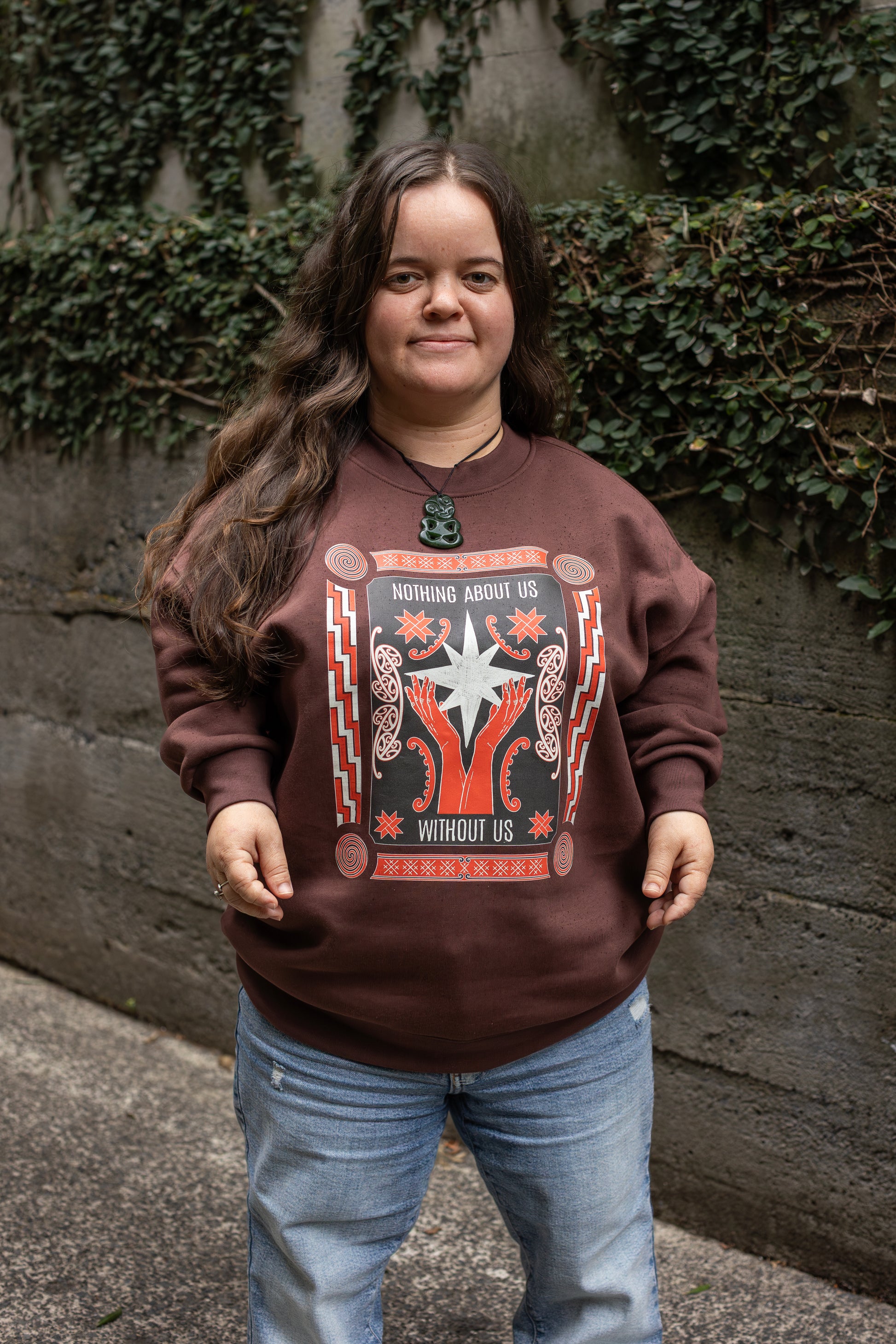 Ella stands in-front of a leafy wall, she wears a brown sweatshirt with a bold design reading "Nothing About Us Without Us." The graphic features red hands, stars, koru and spirals. 