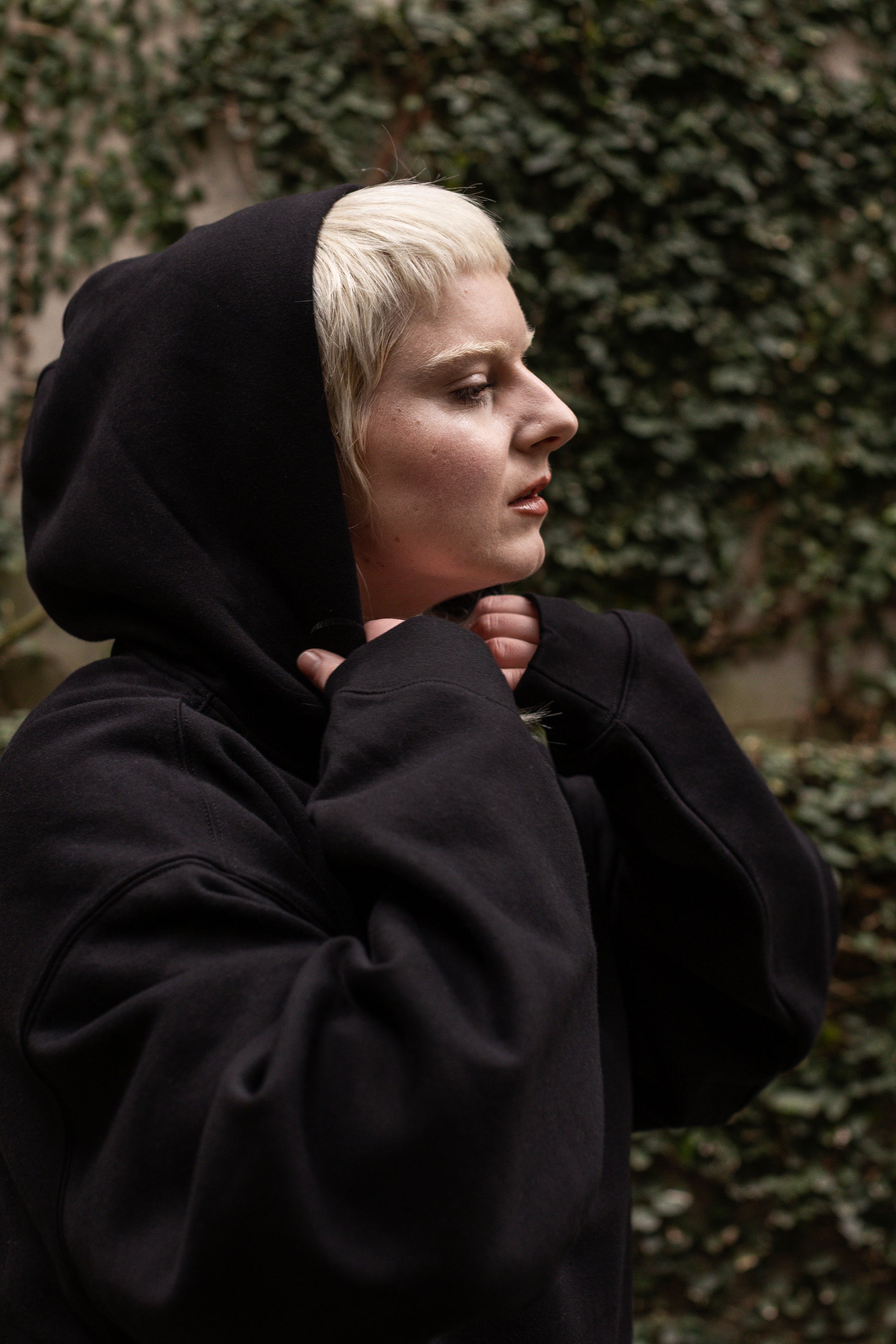 Person wearing a black hoodie, pulling up the hood against a green leafy background