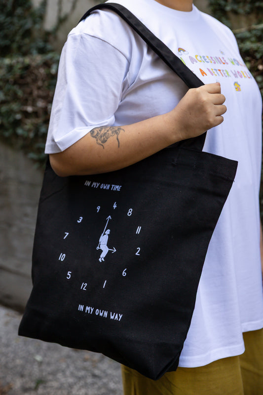 A person wearing a white t-shirt holds a black tote bag with a light blue print. The design features an analogue clock, with jumbled numbers and a person sitting on the clock hands. Above the design reads "IN MY OWN TIME" and below reads "IN MY OWN WAY".
