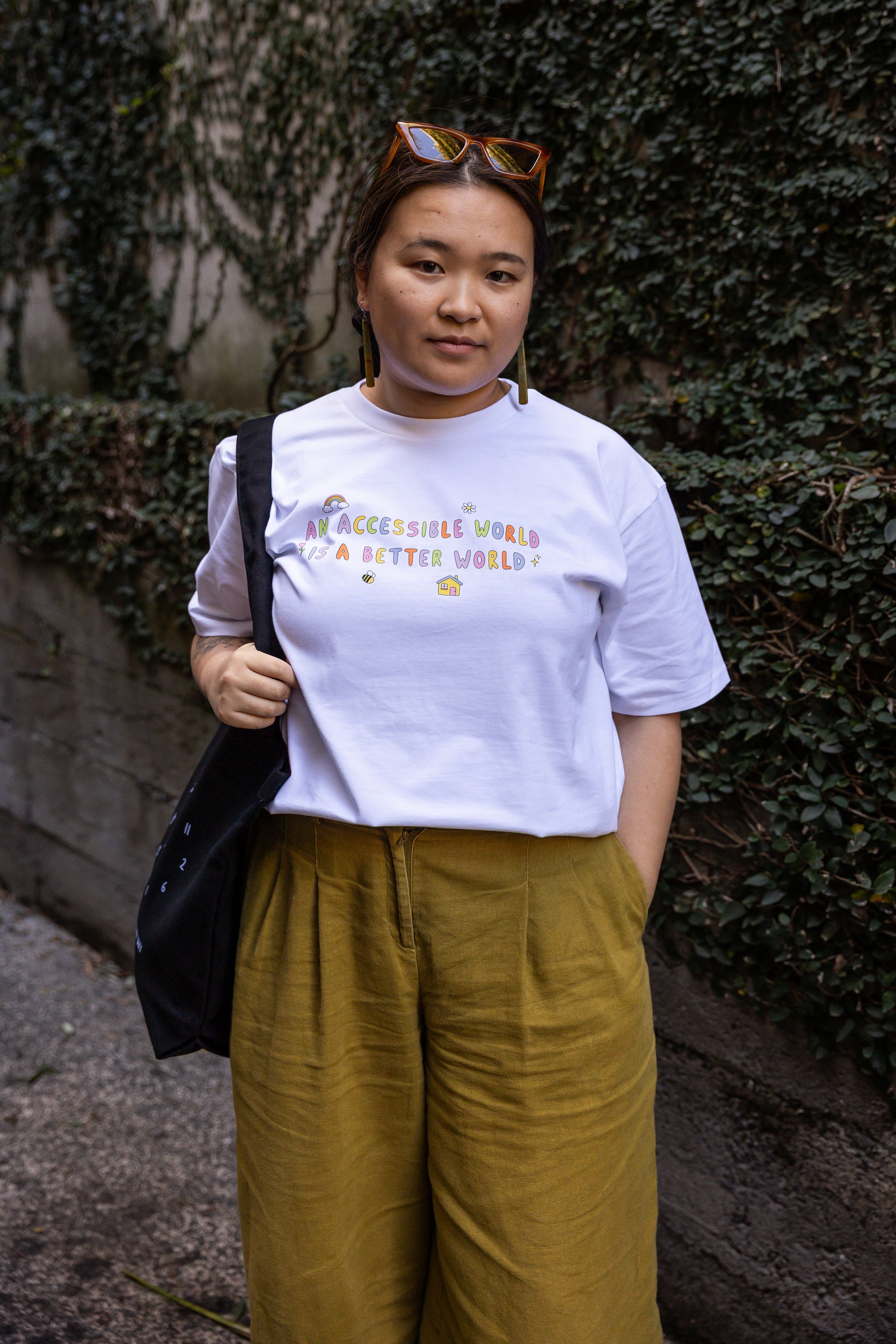 Eda stands in an alley with ivy-covered walls, wearing a white t-shirt with the text "An Accessible World is a Better World" and mustard trousers. She has a relaxed expression, holding a black bag over one shoulder.