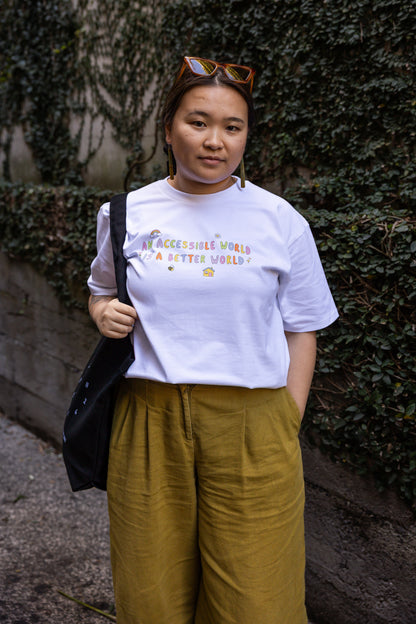 Eda stands in an alley with ivy-covered walls, wearing a white t-shirt with the text "An Accessible World is a Better World" and mustard trousers. She has a relaxed expression, holding a black bag over one shoulder.