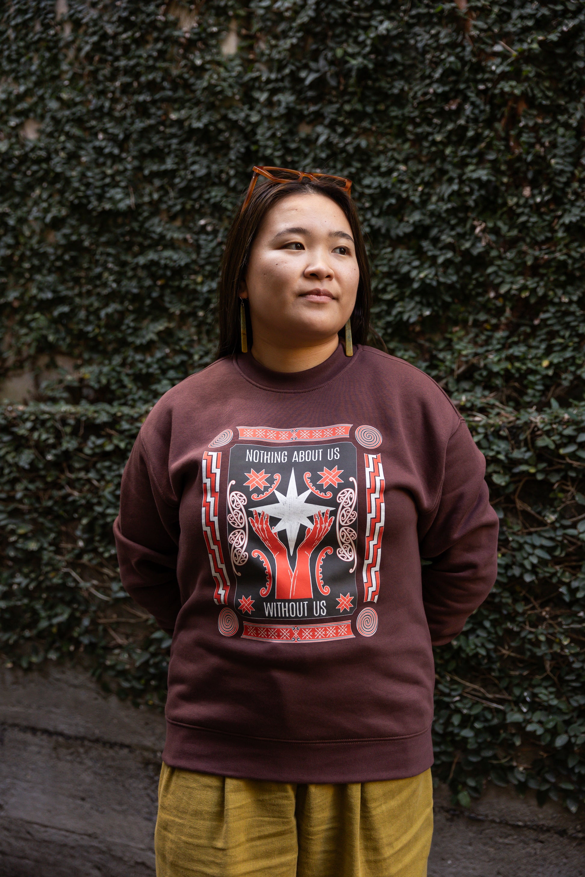Eda stands against a leafy wall, wearing a dark sweatshirt with the phrase "Nothing About Us Without Us." The graphic features red hands, stars, koru and spirals.