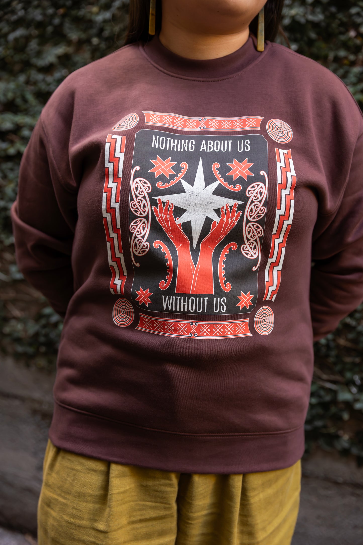 Eda wears a brown sweatshirt with a bold design reading "Nothing About Us Without Us." The graphic features red hands, stars, koru and spirals.