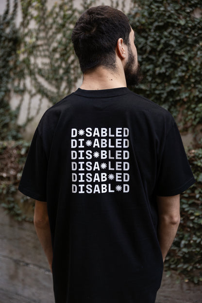 Will stands facing away from the camera, wearing a black T-shirt with "DISABLED" printed multiple times. The white print on the back reads "DISABLED" repeated six times with a tohuwhetū replacing different letters from left to right on each line. He is facing a vine-covered wall.