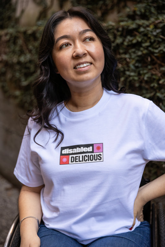 Olivia smiles warmly, wearing a white T-shirt that reads "disabled DELICIOUS." The design features two tohuwhetū and is in pink, red, black and white.