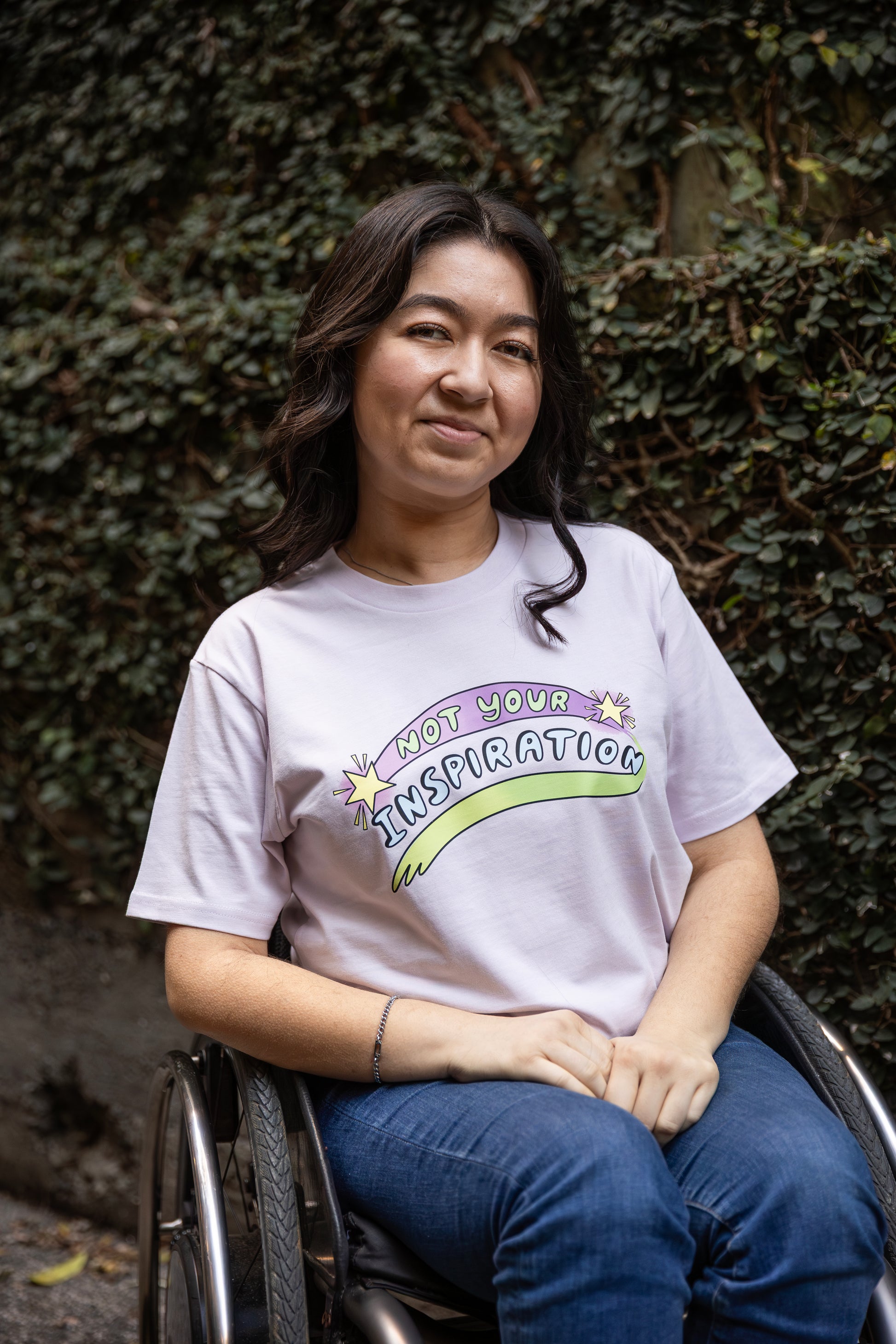 Olivia wearing a light pink T-shirt with the phrase "Not Your Inspiration" in colourful letters sits in her wheelchair against a leafy background. The words 'Not your' appear in lime green on a purple swoosh, positioned between two yellow stars. Beneath these words, 'inspiration' appears in all caps, while a second stroke of colour, which appears like a green-yellow paint stroke, provides a boundary of support at the base of the print. 