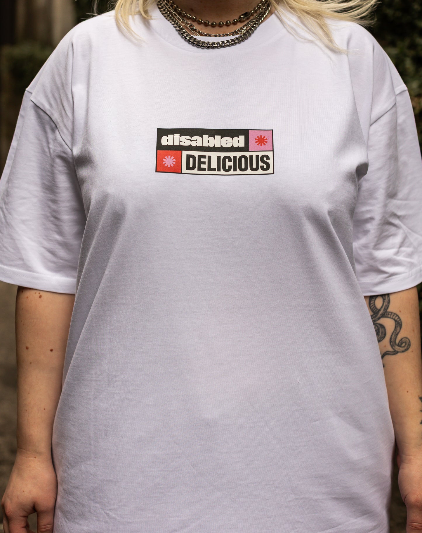 A close up of Von's torso wearing a white T-shirt that reads "disabled DELICIOUS." The design features two tohuwhetū and is in pink, red, black and white.