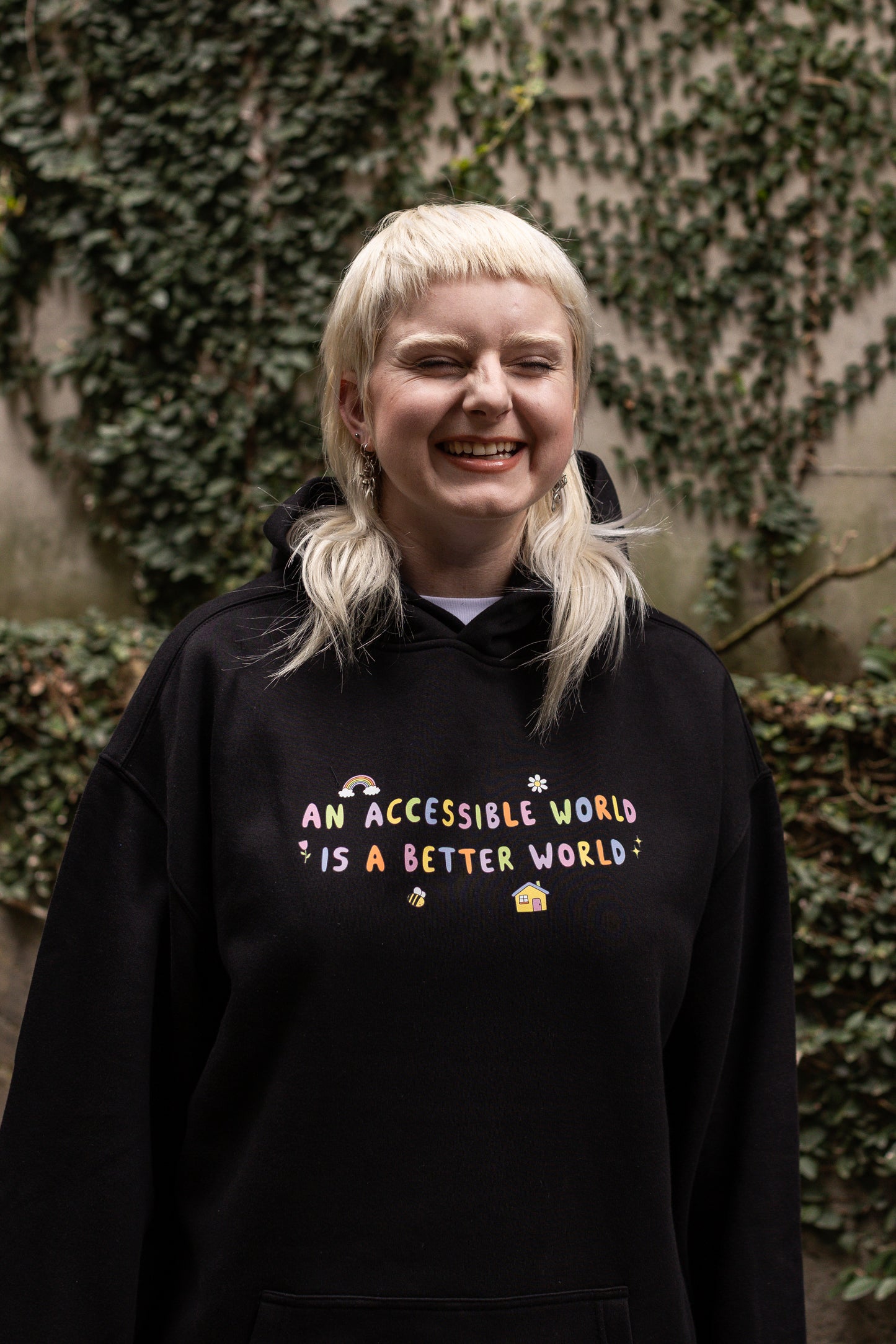 Von wears a black hoodie with colourful text which reads "An accessible world is a better world". The design has small designs around it including flowers, a rainbow, a bee, a house and a star.