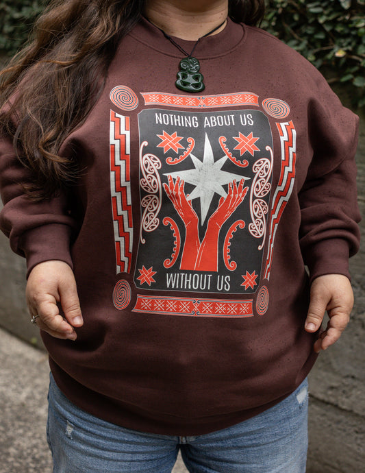Ella wears a brown sweatshirt with a bold design reading "Nothing About Us Without Us." The graphic features red hands, stars, koru and spirals.